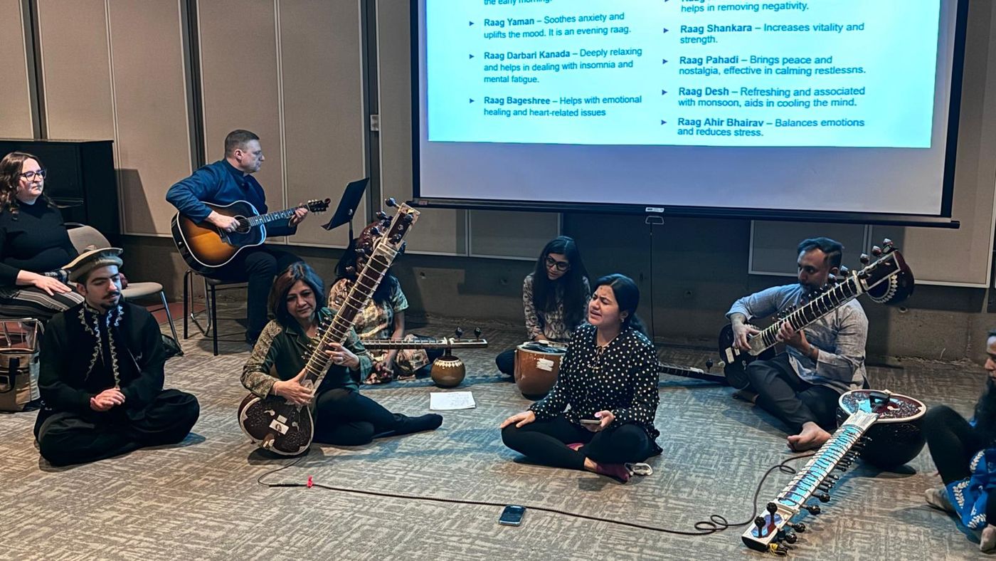 Sitar and guitar players seated on the floor in front of a projected list of ragas